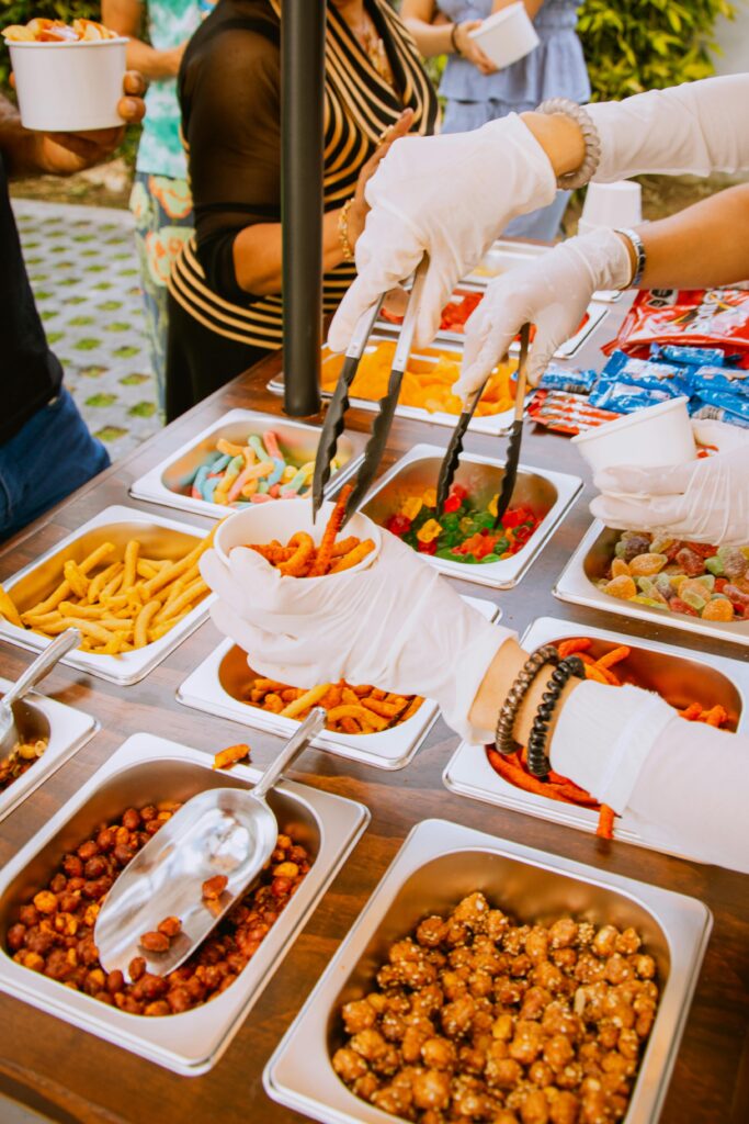 Gloved hands serving at an outdoor snack buffet with a variety of finger foods.